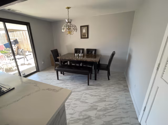 A dining area with a rectangular marble-top table surrounded by four black chairs and a black bench. The floor has white tiles with gray marble patterns. A sliding glass door leads to an outdoor patio with a wooden fence and some outdoor furniture. A decorative hanging light fixture is above the table, and a small framed picture is on the wall.