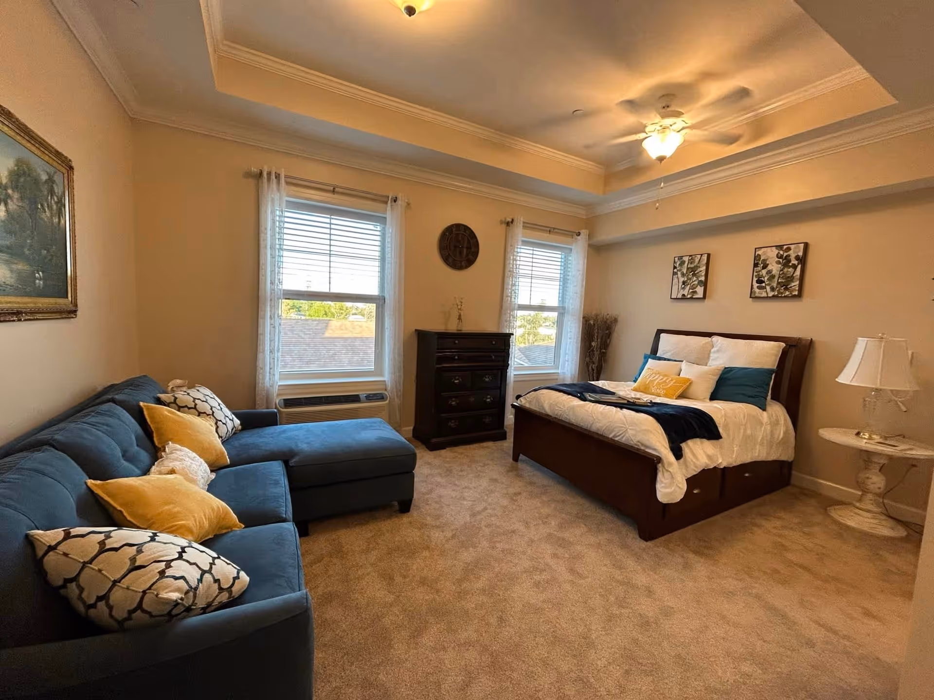 A cozy bedroom in a senior living facility featuring a large bed with white and blue bedding, a blue sectional sofa with patterned and yellow pillows, two windows with white curtains, a dark wooden dresser, a round side table with a lamp, and framed artwork on the walls.