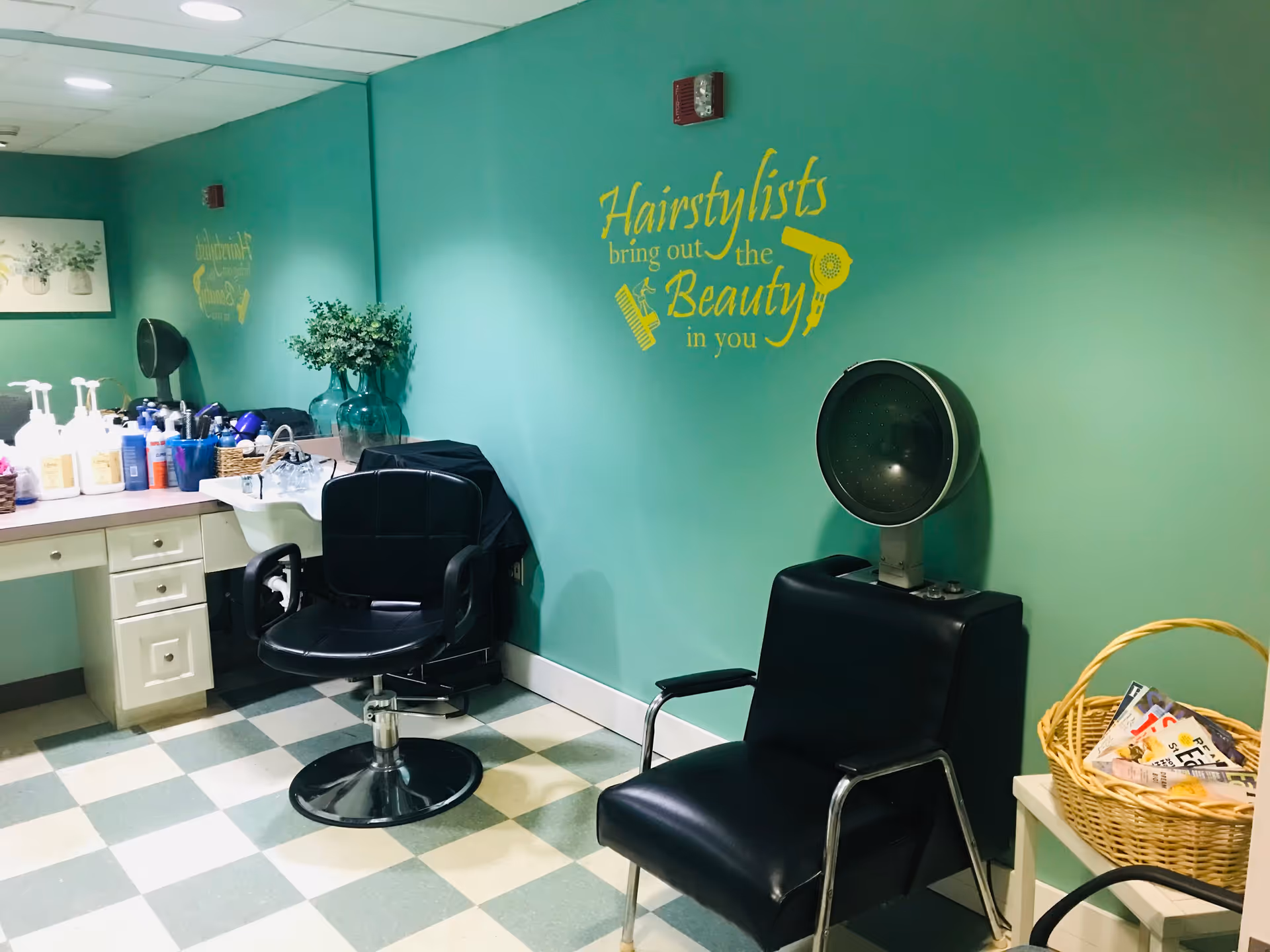Interior of a senior living community hair salon with teal walls, a black salon chair in front of a sink, a black hair dryer chair, a basket with magazines on a small table, and a wall decal that reads 'Hairstylists bring out the Beauty in you'.