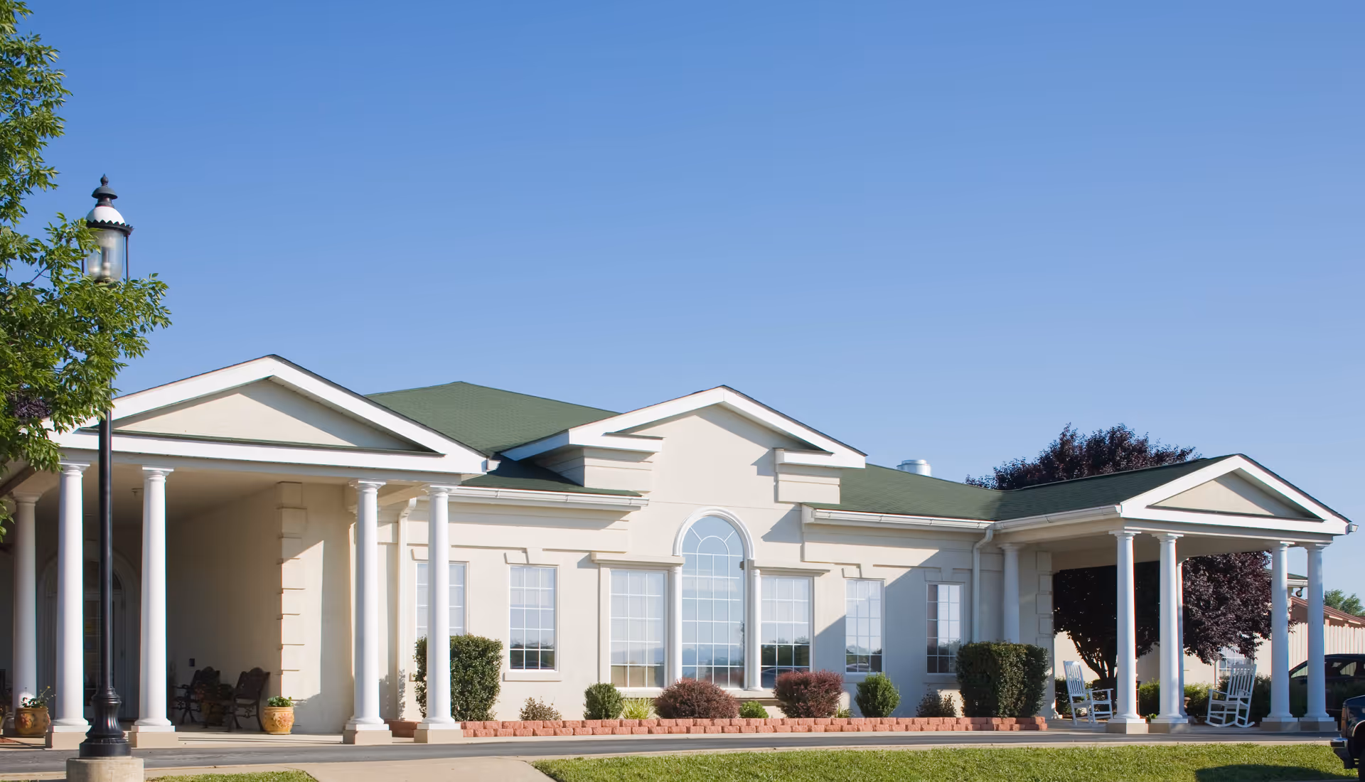 Front exterior of a light-colored single-story building with white columns, a green roof, an arched central window, and a covered porch with rocking chairs.