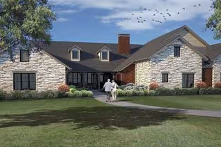 Front exterior view of a single-story assisted living facility with stone and brick facade, a dark shingled roof, and a chimney. A couple is walking on a paved path towards the entrance. The sky is blue with some clouds and birds flying.