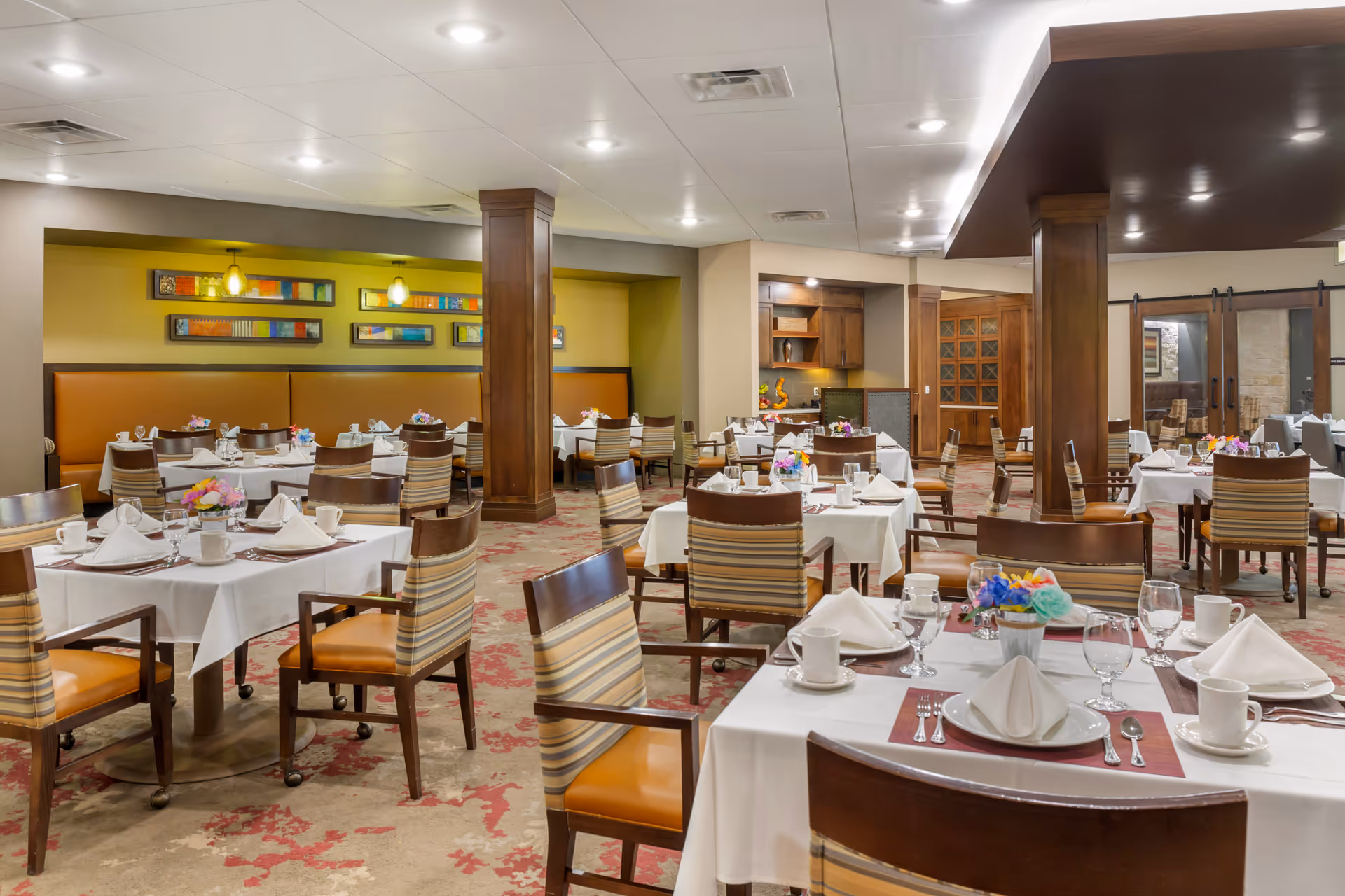 A well-lit dining room with multiple tables covered in white tablecloths, each set with plates, napkins, cups, glasses, and silverware. The chairs have striped upholstery with wooden frames. The room features wooden columns, colorful wall art, and a carpet with a red and beige pattern.
