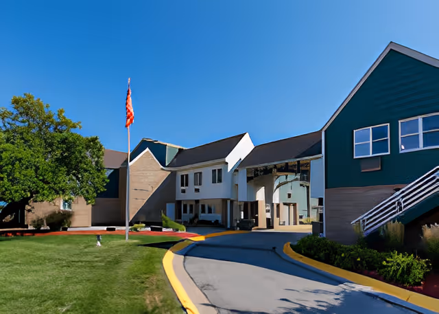 Exterior view of Hickory Villa Assisted Living facility showing a driveway leading to the entrance with a covered drop-off area. The building has green and beige siding with multiple windows, a flagpole with an American flag, and landscaped greenery including a large tree and bushes.