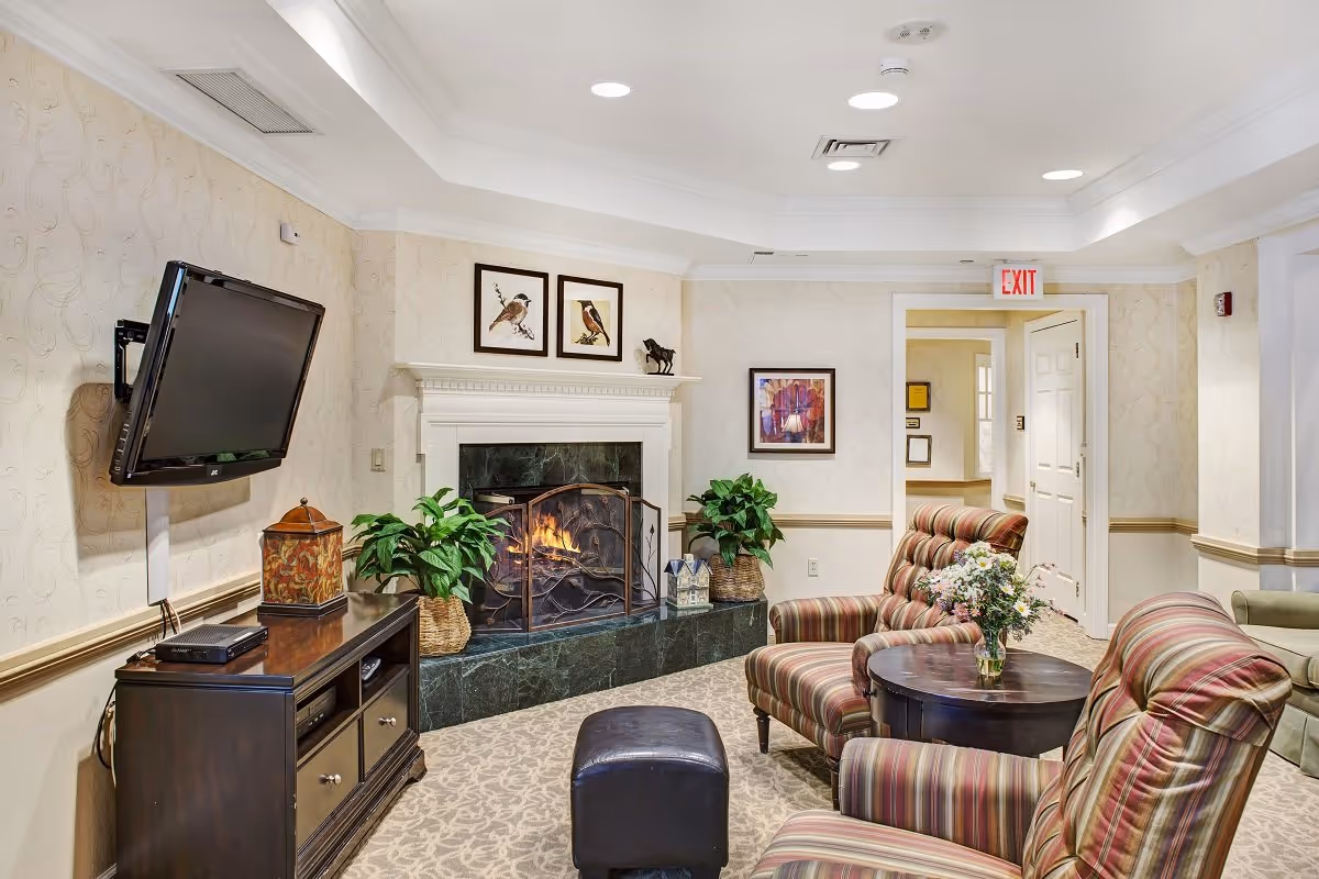 A cozy living room area with a lit fireplace surrounded by green marble. Two striped armchairs and a small round wooden table with a vase of flowers are in the foreground. A flat-screen TV is mounted on the wall above a dark wooden cabinet. The walls are light-colored with subtle patterns, decorated with framed pictures and plants in woven baskets. A doorway with an exit sign is visible in the background.