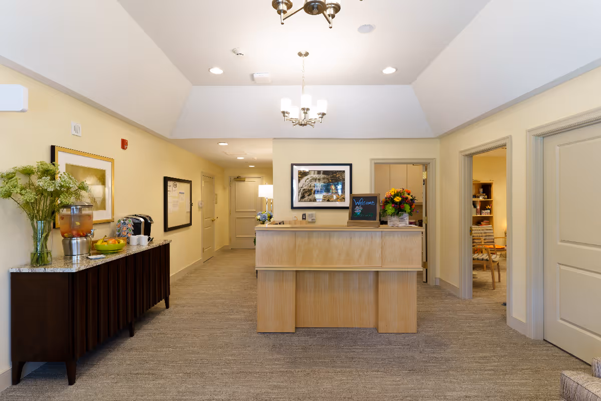 Reception area in a senior living facility with a wooden front desk, a welcome sign, a bouquet of flowers, and a framed picture on the wall. To the left, there is a sideboard with a beverage dispenser, cups, a coffee maker, and a bowl of fruit. The area is well-lit with ceiling lights and a chandelier, and there are doors leading to other rooms.