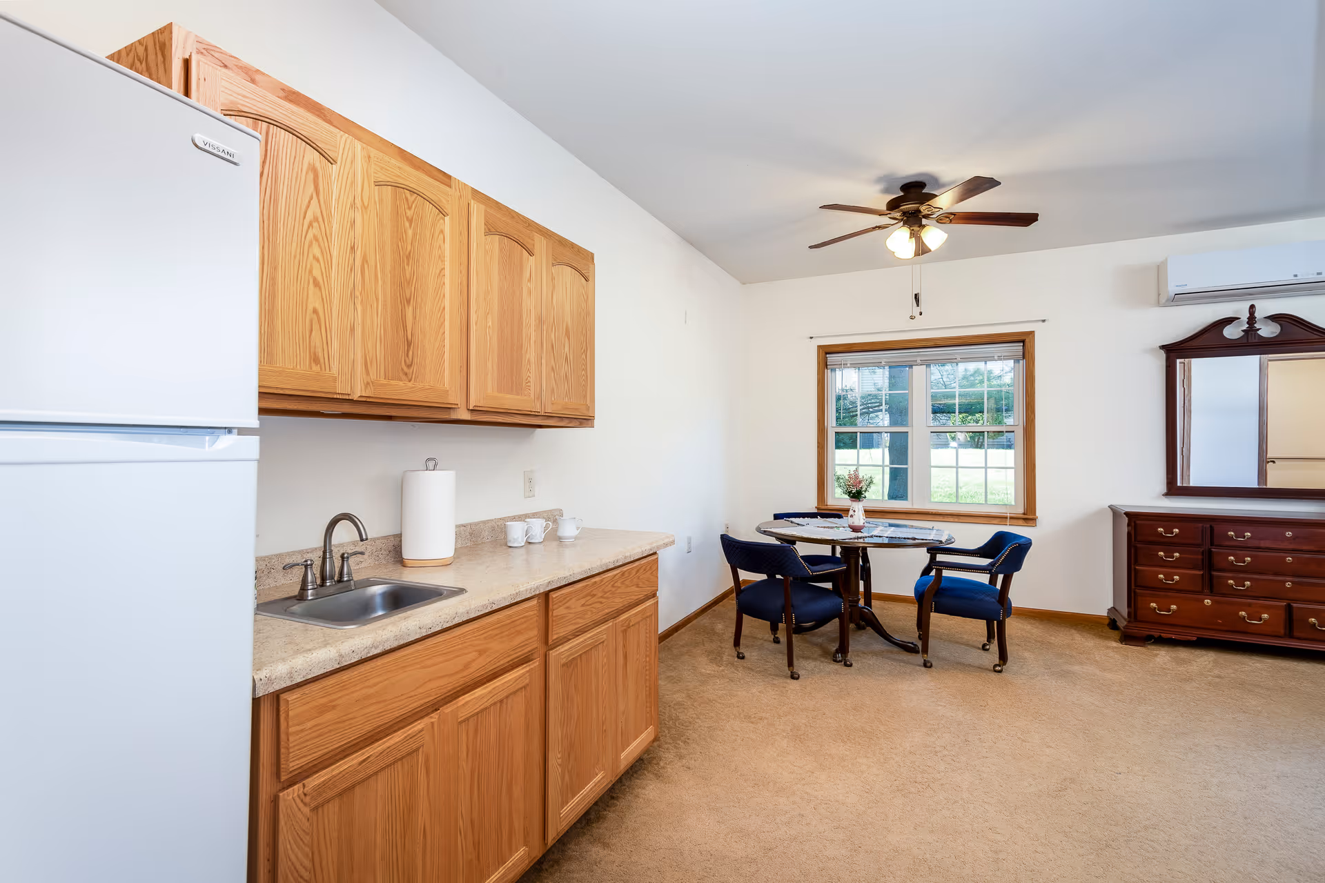 Small senior living kitchen and dining area with oak cabinets, a sink and refrigerator on the left and a round table with blue chairs by a window.