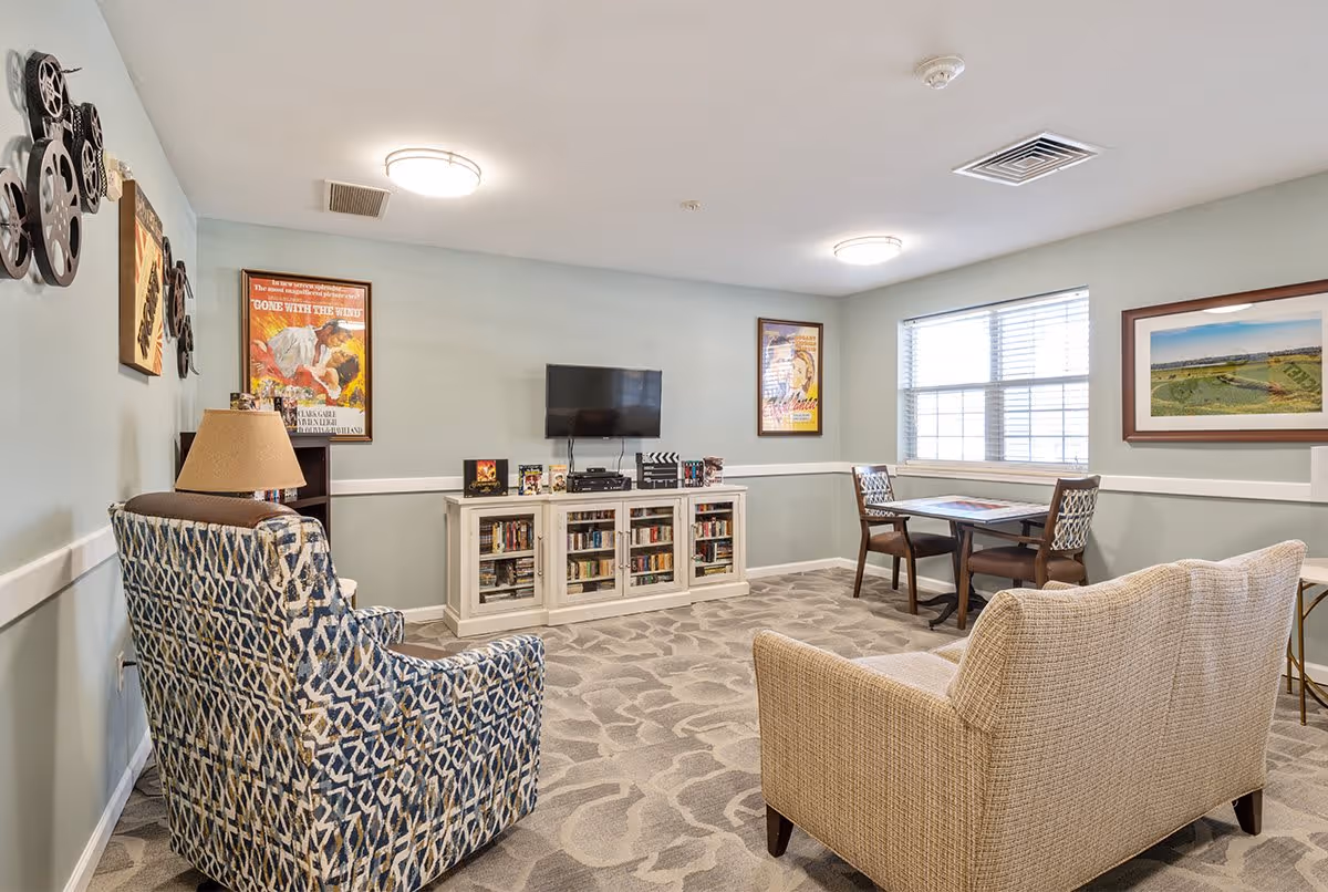 A cozy senior living common area with a patterned armchair, a beige loveseat, a small table with two chairs near a window, a white cabinet with books and a flat-screen TV mounted above it, and movie-themed wall decorations including framed posters and film reels.