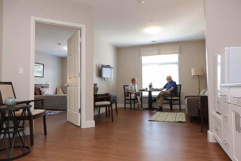 Two seniors sit at a small table in a bright assisted-living living room with seating, a wall TV, and an adjacent open bedroom.