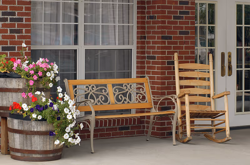 Outdoor seating area with a wooden bench and a wooden rocking chair on a concrete porch in front of a brick wall with windows and glass doors. Two large wooden barrels filled with colorful flowers are placed next to the bench.