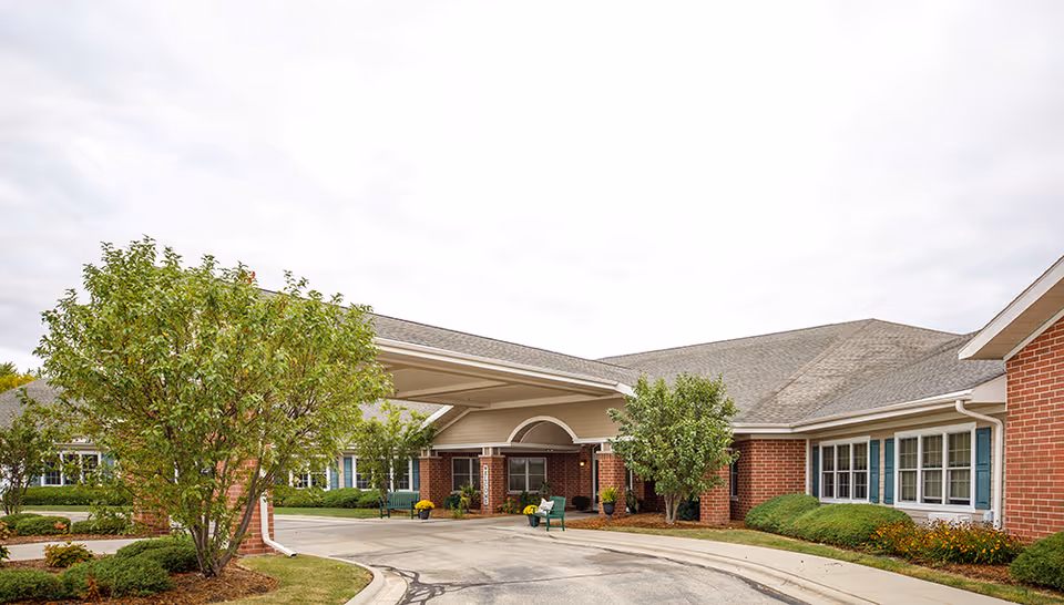 Exterior view of Parkside Manor Assisted Living and Memory Care building with a covered entrance, brick walls, multiple windows, and landscaped greenery including trees and bushes along the driveway.