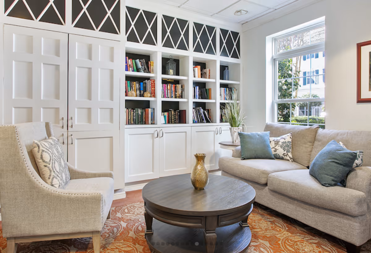 A cozy living room with a beige armchair and a gray sofa adorned with blue and patterned pillows. There is a round wooden coffee table with a decorative vase on it, placed on a patterned rug. Behind the seating area is a white built-in bookshelf filled with books and decorative items. A large window lets in natural light, and a framed picture hangs on the wall.
