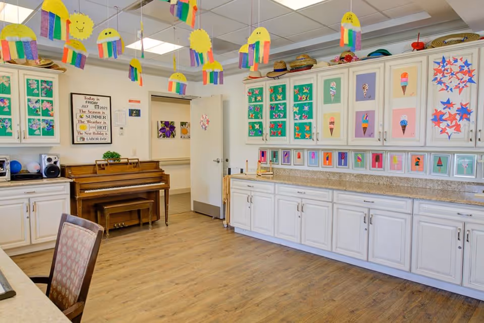 Bright activity room with a piano, long white cabinets and countertop, colorful children's artwork on the walls, and hanging paper decorations.
