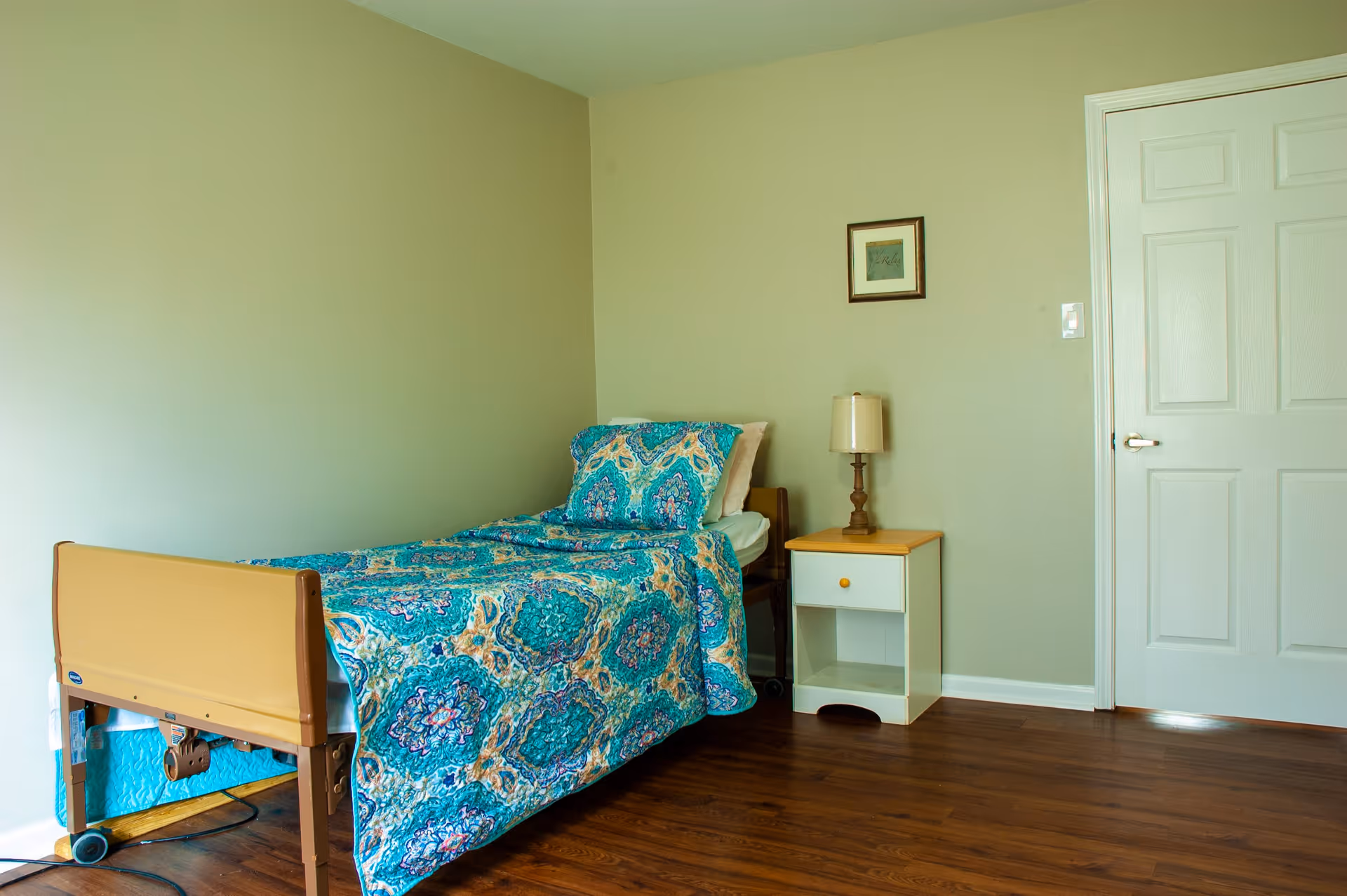A simple bedroom with a single bed covered in a blue and green patterned quilt and pillow. Next to the bed is a small white nightstand with a wooden top and a lamp. A framed picture hangs on the beige wall above the nightstand. The room has wooden flooring and a closed white door.