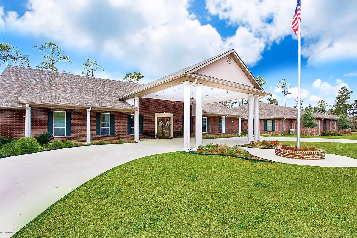 Exterior view of a single-story brick building with a covered entrance supported by white columns, a curved driveway, a well-maintained lawn, and an American flag on a flagpole under a partly cloudy blue sky.
