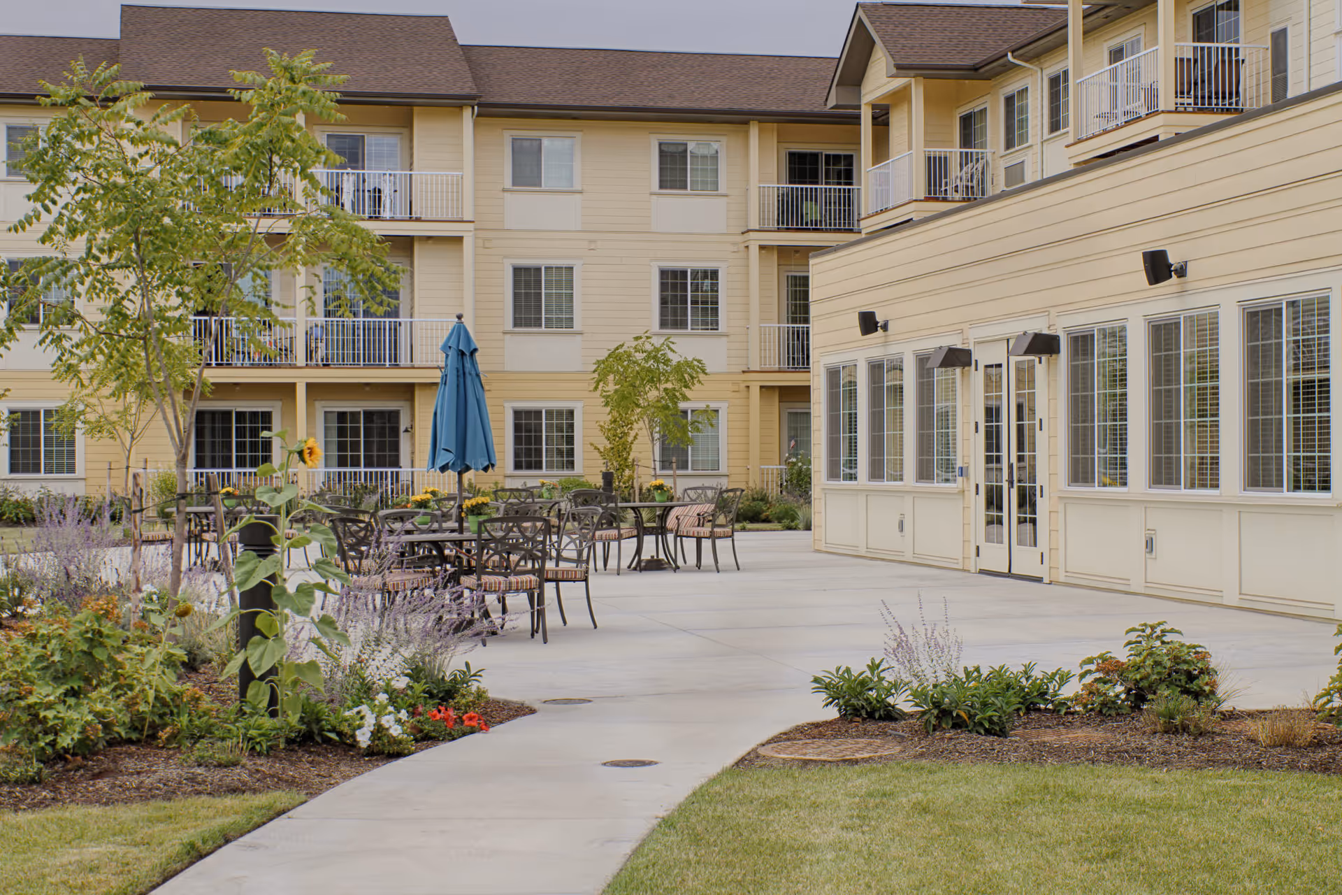 Outdoor patio area at Volante Senior Living of Eugene featuring several tables and chairs with a blue umbrella, surrounded by landscaped garden beds and trees, adjacent to a multi-story beige building with balconies and large windows.