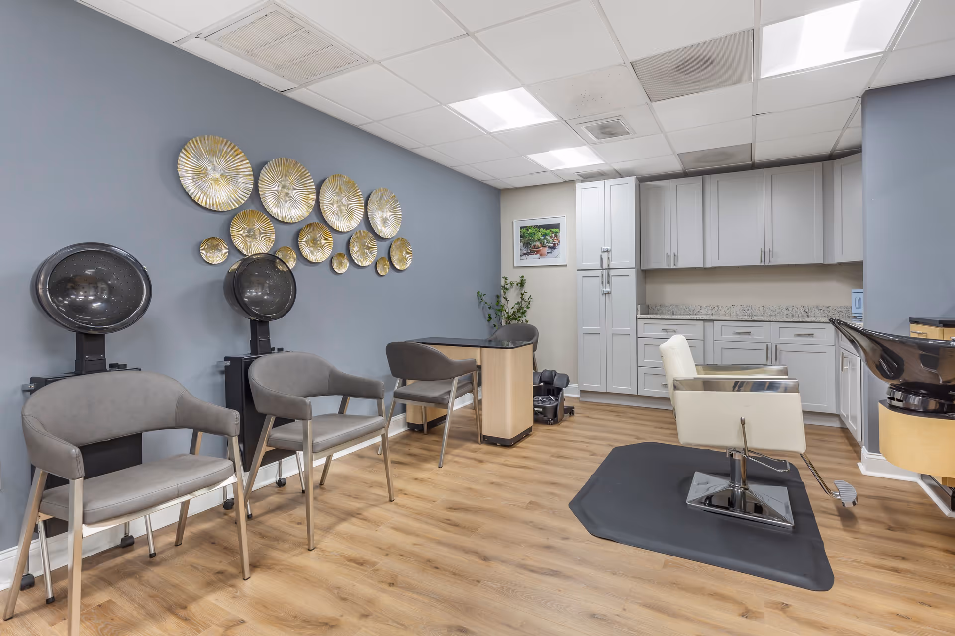 Interior view of a salon area in a retirement community featuring two hair drying chairs with hooded dryers, a styling chair on a black mat, a small table with two chairs, and white cabinetry along the back wall. The walls are painted blue and beige, with decorative gold circular wall art and a framed picture. The floor is wood laminate.