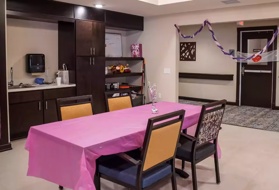 Communal dining/activity room with a pink-covered table, chairs, and a small kitchenette and storage cabinets along the wall.
