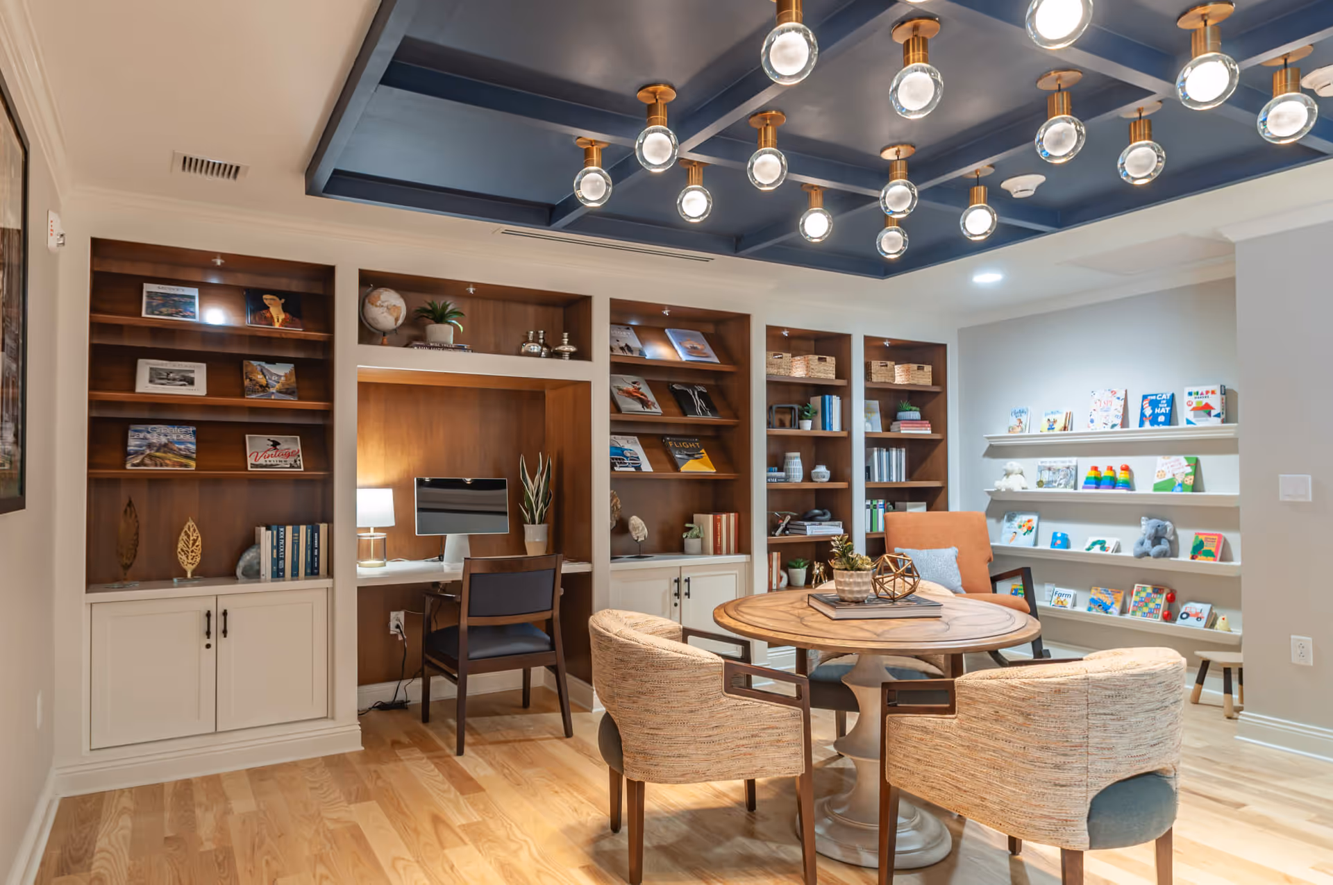 A cozy interior room featuring a round wooden table surrounded by four upholstered chairs. Behind the table is a built-in wooden shelving unit with books, decorative items, and a computer workstation with a chair. To the right, there are wall-mounted shelves displaying children's books and toys. The ceiling has a blue coffered design with multiple modern globe light fixtures. The floor is light wood.