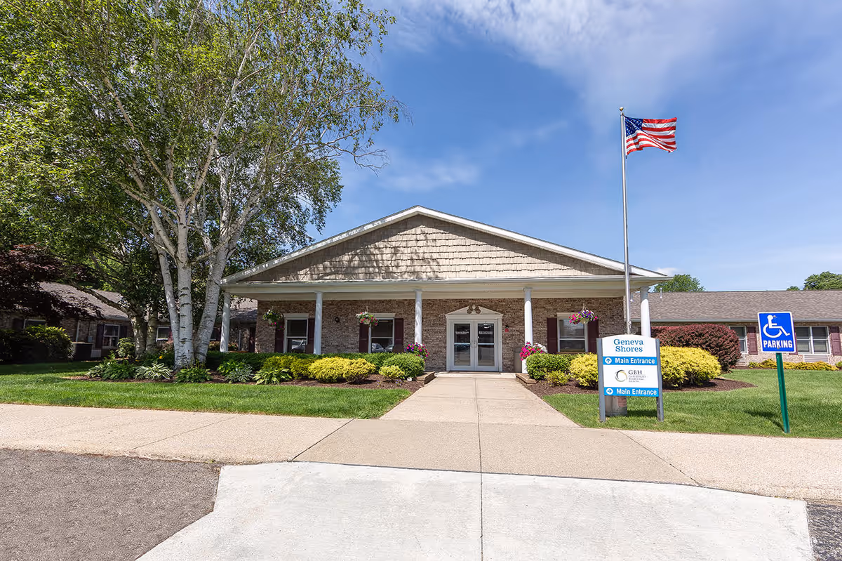 Front entrance of Geneva Shores nursing facility with an American flag, walkway, landscaping, and a handicap parking sign.