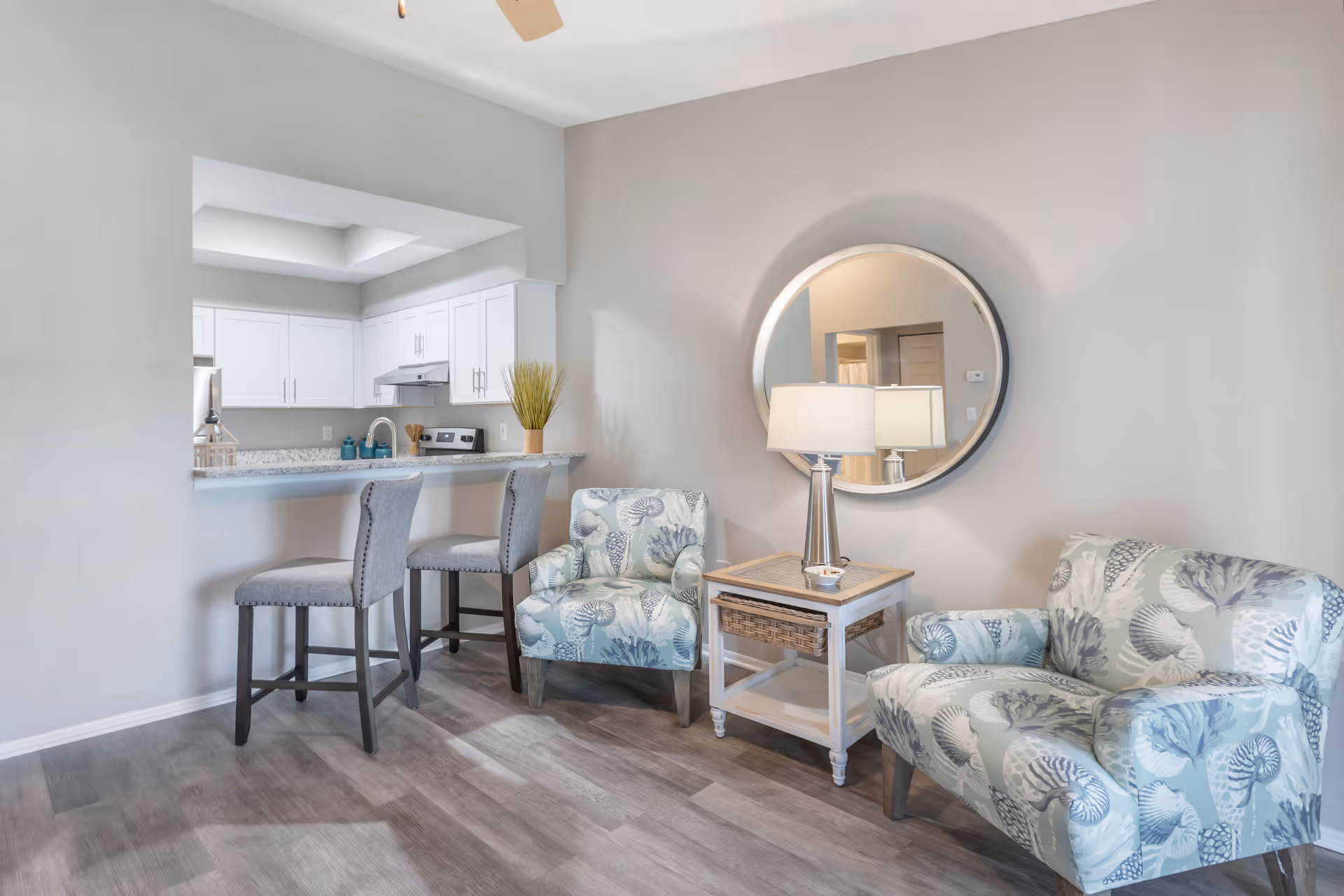 Cozy sitting area with two patterned armchairs, a side table and lamp beneath a round mirror, adjacent to a kitchen counter with bar stools.