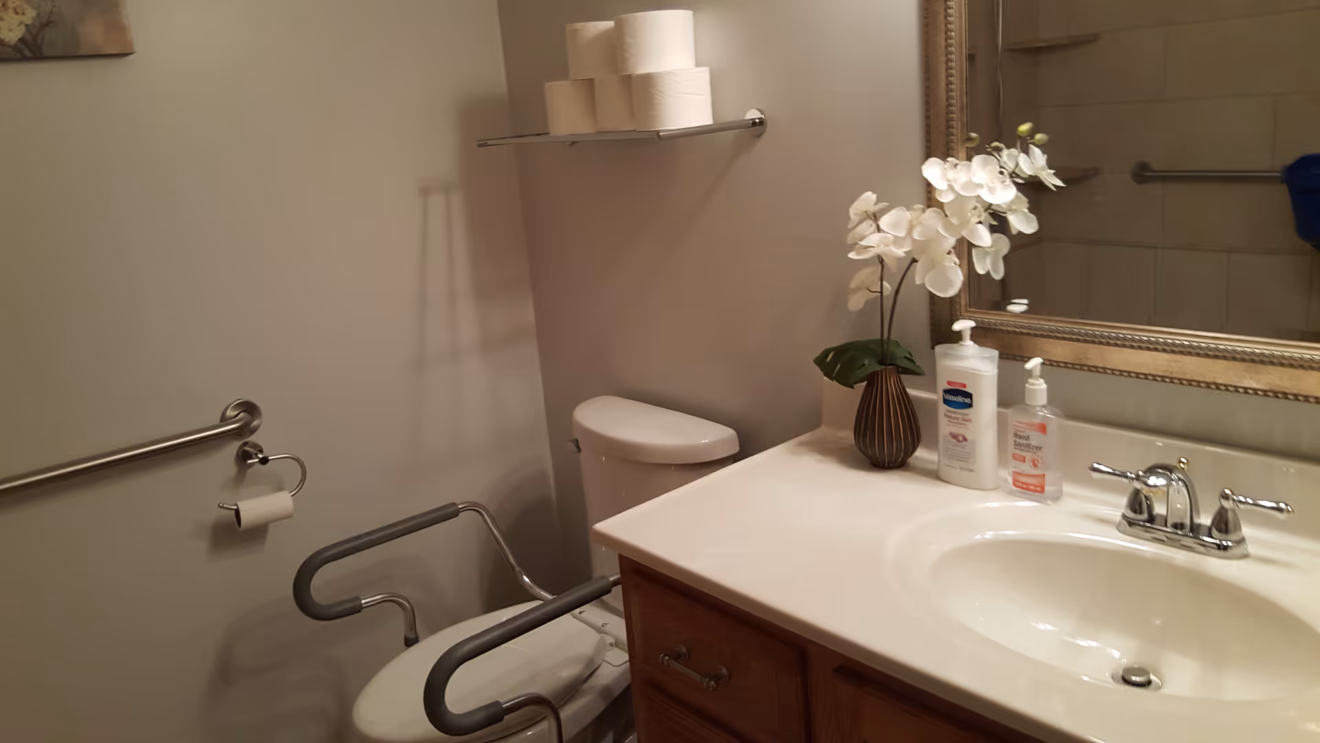 A clean bathroom with a toilet equipped with safety rails, a toilet paper holder with a roll, a shelf holding extra toilet paper rolls, a sink with a faucet, a large mirror, a vase with white flowers, and two bottles of hand soap and lotion on the countertop.