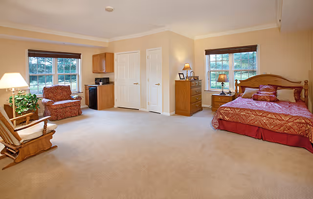 A spacious bedroom with beige walls and carpet. The room features a wooden bed with red bedding and pillows, a wooden dresser with a lamp and picture frame, a wooden nightstand with a lamp, a red upholstered armchair, a wooden rocking chair, a small kitchenette area with a microwave and mini fridge, and two windows with brown blinds letting in natural light.