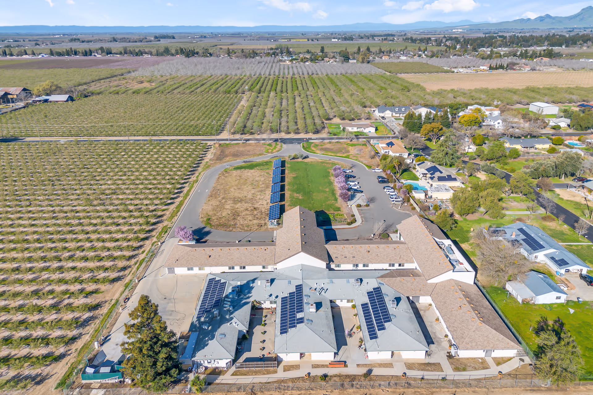 Aerial view of a single-story senior living complex with solar panels on the roofs, a parking area, and surrounding orchards and homes.