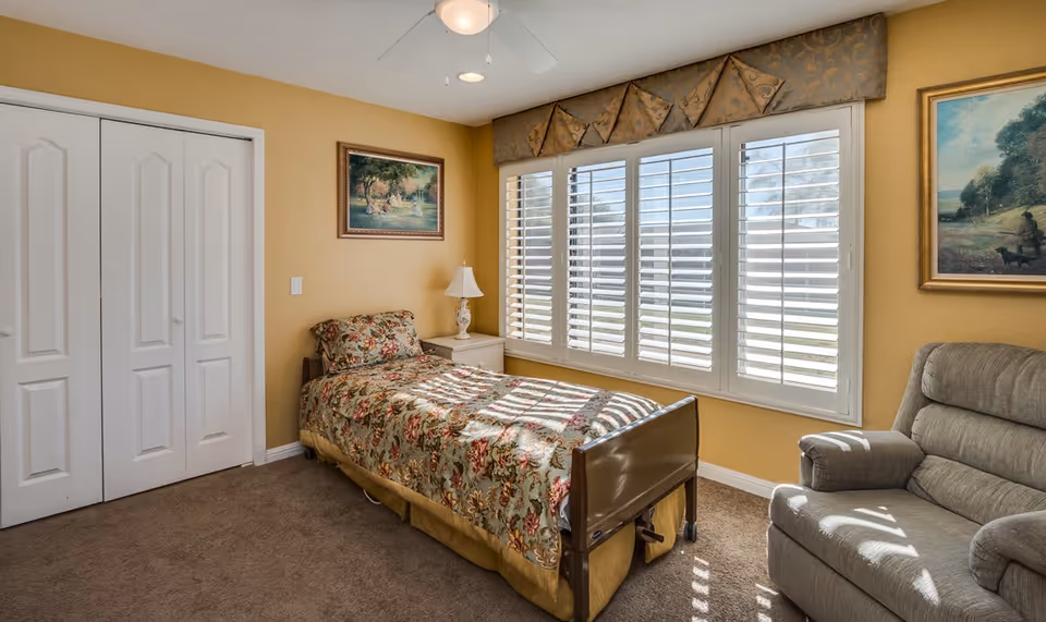 A cozy bedroom with a single bed covered in a floral bedspread, a nightstand with a lamp, a gray armchair, two framed paintings on yellow walls, a large window with white plantation shutters, and a ceiling fan with a light.