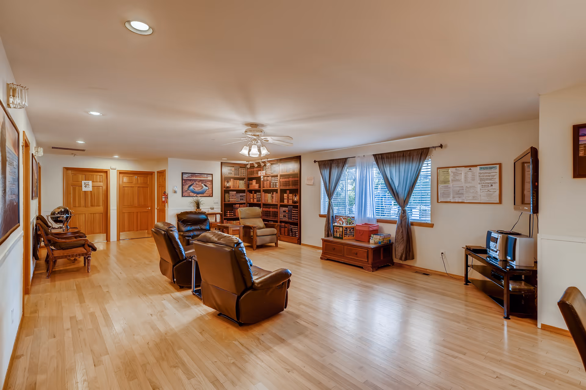 A spacious living room area with wooden flooring, three brown leather recliners, a beige armchair, and a wooden chest under a window with blue curtains. The room has a ceiling fan with lights, framed pictures on the walls, a bookshelf filled with books, and a TV mounted on the wall above a black stand with a stereo system.