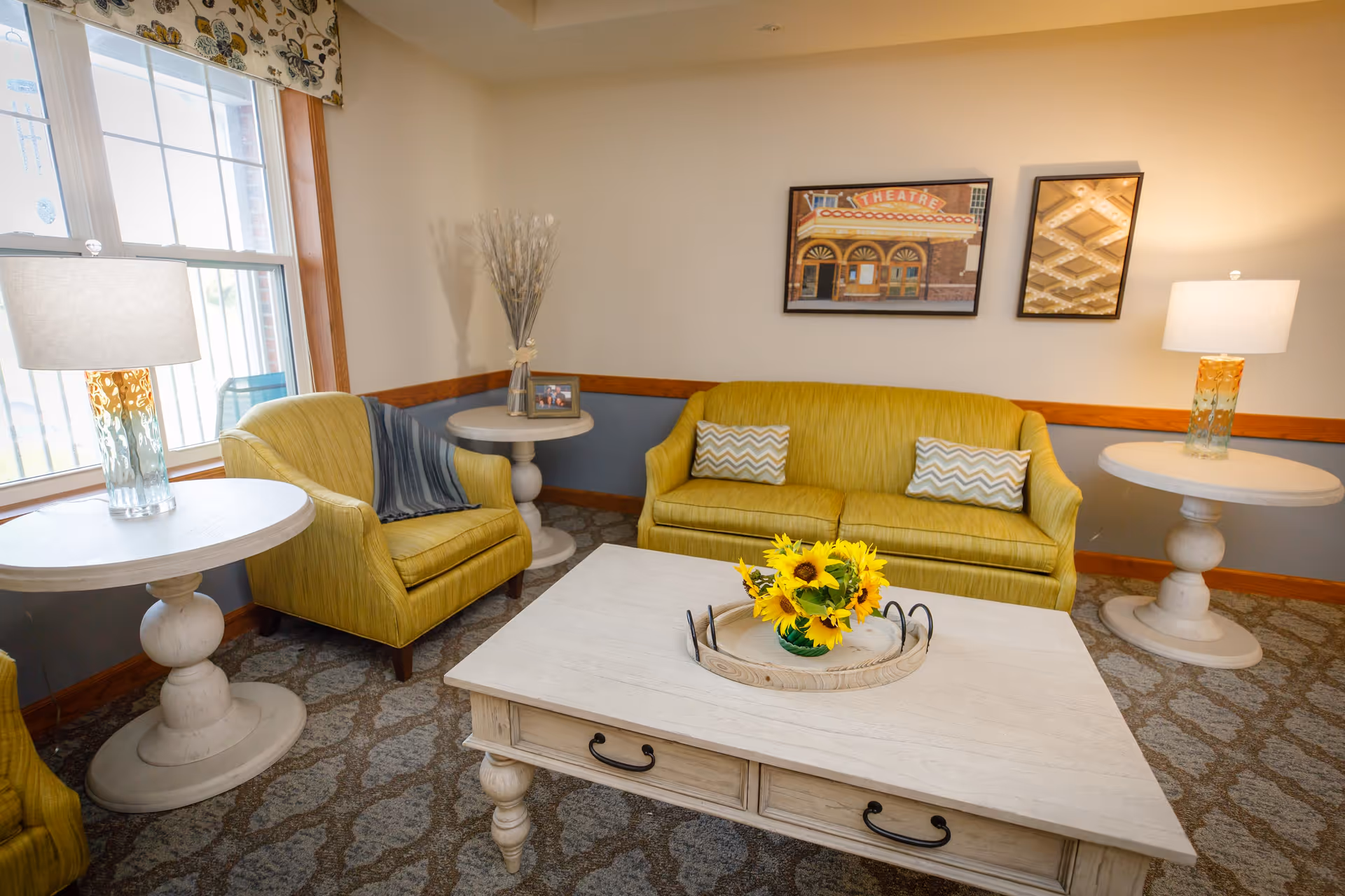 A cozy living room with a yellow sofa and matching armchair, two round white side tables with lamps, a white coffee table with a tray holding a vase of sunflowers, a window with floral curtains, and two framed pictures on the wall.