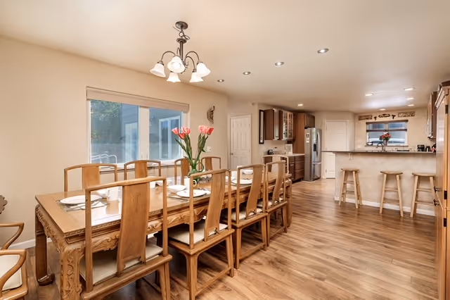 Open-plan dining area with a long wooden table set for eight and an adjacent kitchen with a breakfast bar and stools.