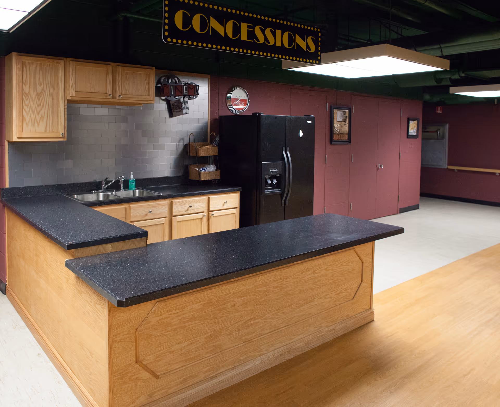 Interior view of a concession stand area with wooden cabinets, black countertops, a double sink, a black refrigerator, and a sign overhead that reads 'CONCESSIONS'. The walls are painted maroon and there are framed pictures hanging on them.
