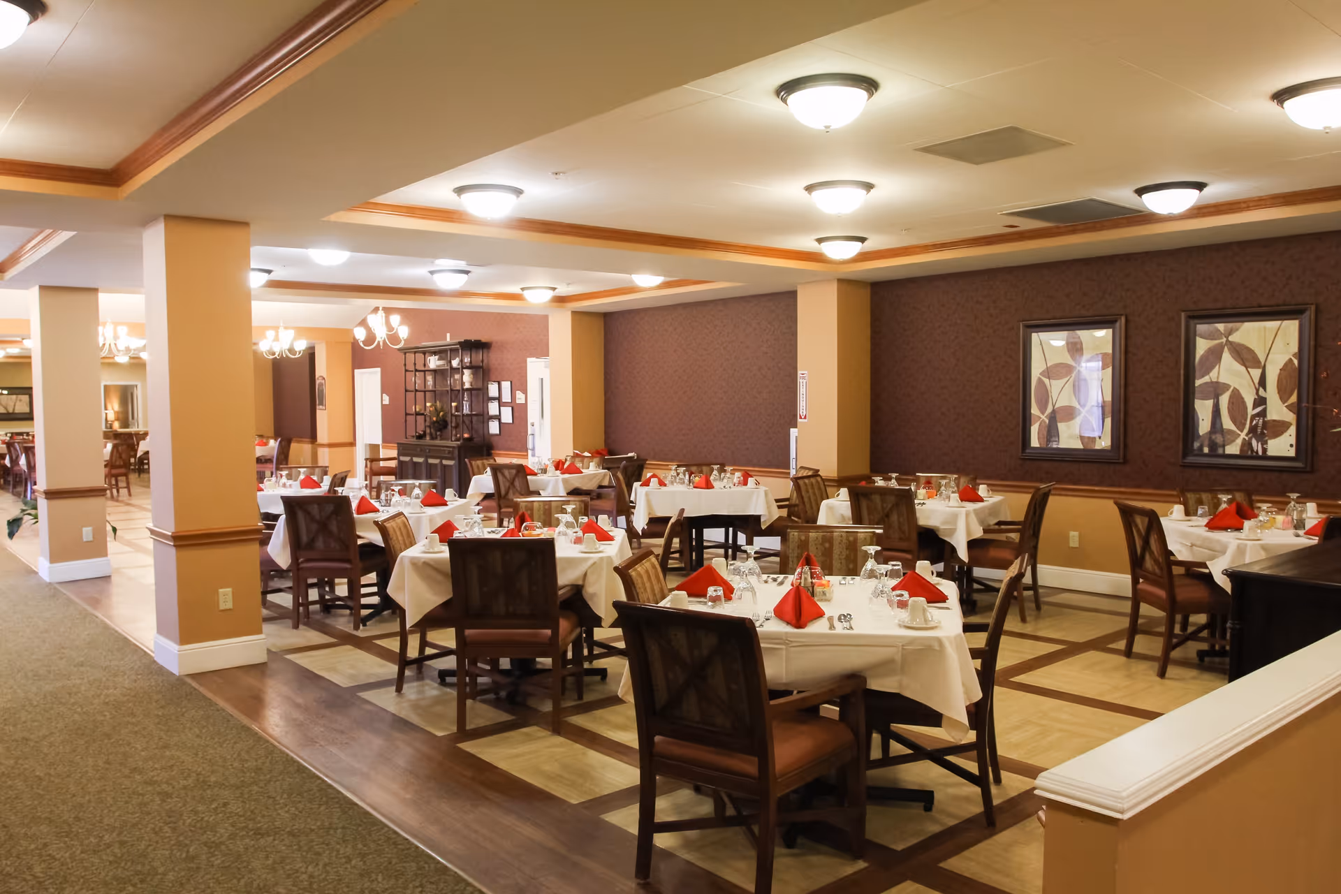 A dining room in an assisted living facility with multiple tables covered in white tablecloths, each set with glasses, plates, and red folded napkins. The room has warm beige and brown tones, framed artwork on the walls, and ceiling lights providing illumination.