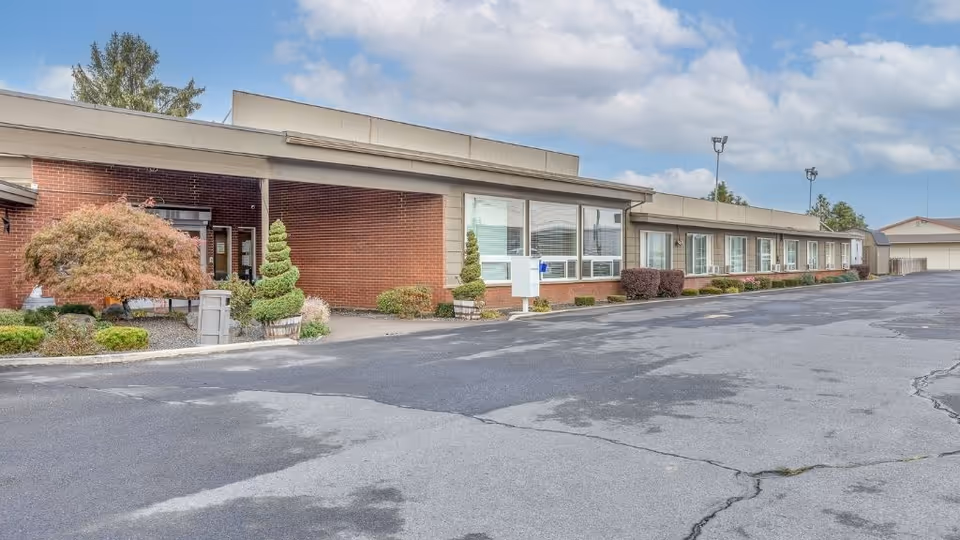 Front exterior of a single-story brick senior living facility with a large asphalt driveway and landscaped shrubs.