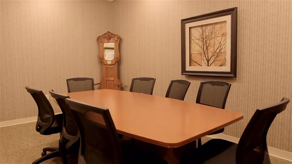 Conference-style room with a long wooden table surrounded by black mesh chairs, a framed picture on the wall and a decorative mirror in the corner.