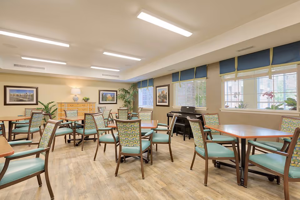 A bright and spacious dining room with multiple wooden tables and chairs featuring light blue cushions. The room has large windows with blue valances, framed pictures on the walls, a wooden dresser with a lamp and plant, and a piano in the corner. The floor is light wood, and the ceiling has recessed lighting.