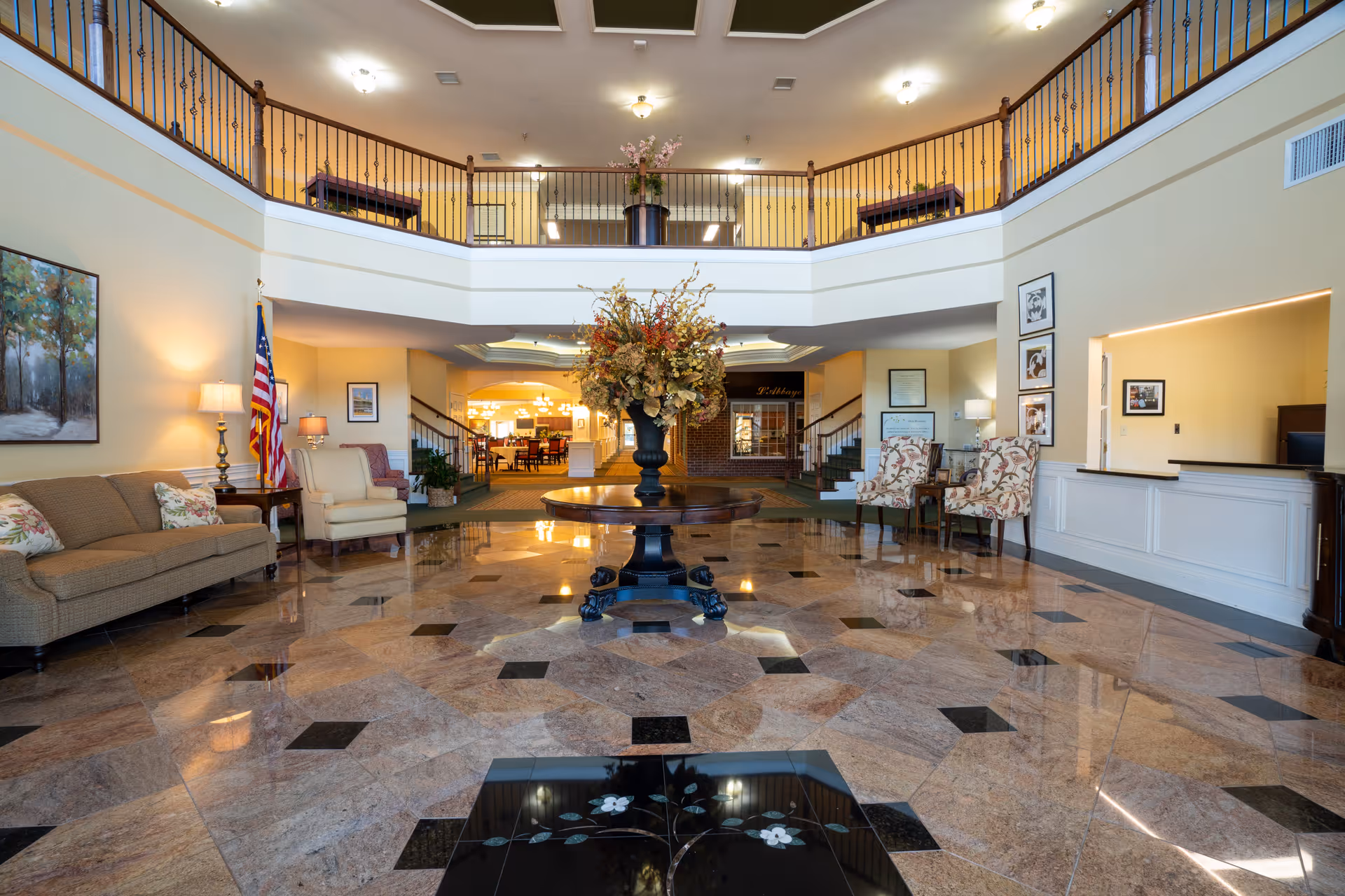 Spacious and well-lit senior living facility lobby with a polished marble floor featuring a floral design, a round wooden table with a large flower arrangement in the center, comfortable seating including sofas and armchairs, an American flag, framed artwork on the walls, and a balcony overlooking the area.