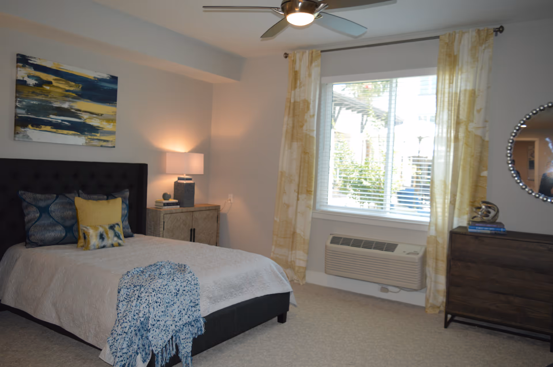 A bedroom with a neatly made bed featuring a white quilt, blue and yellow decorative pillows, and a blue throw blanket. There is a nightstand with a lamp and books next to the bed, a window with yellow patterned curtains, a wall-mounted air conditioning unit below the window, and a dresser with a round mirror and decorative items on top.