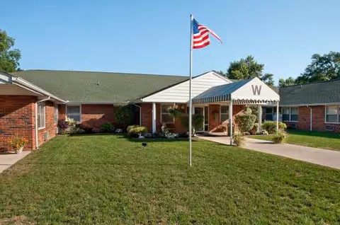 Brick senior living building with green lawn, walkway, and an American flag at entrance.