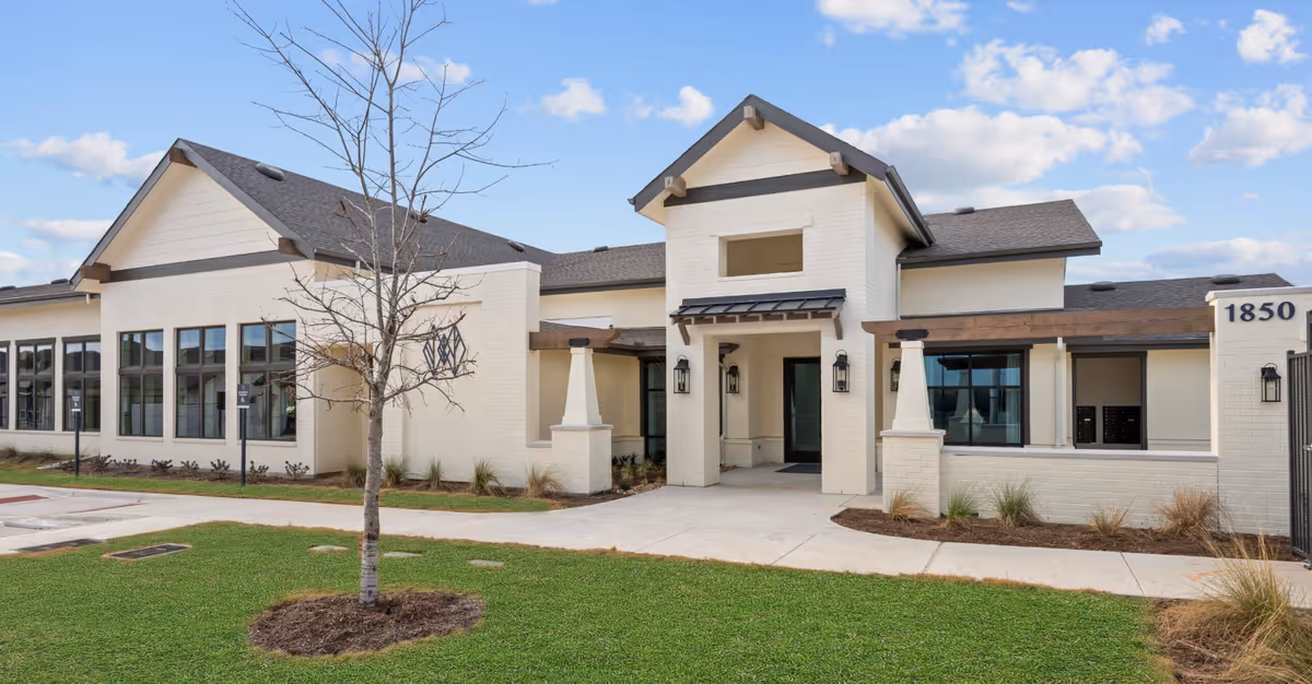 Exterior view of Woodland Cottages Senior Living College Station building with a modern design, light-colored brick walls, large windows, and a covered entrance. There is a small tree and green lawn in front under a partly cloudy sky.