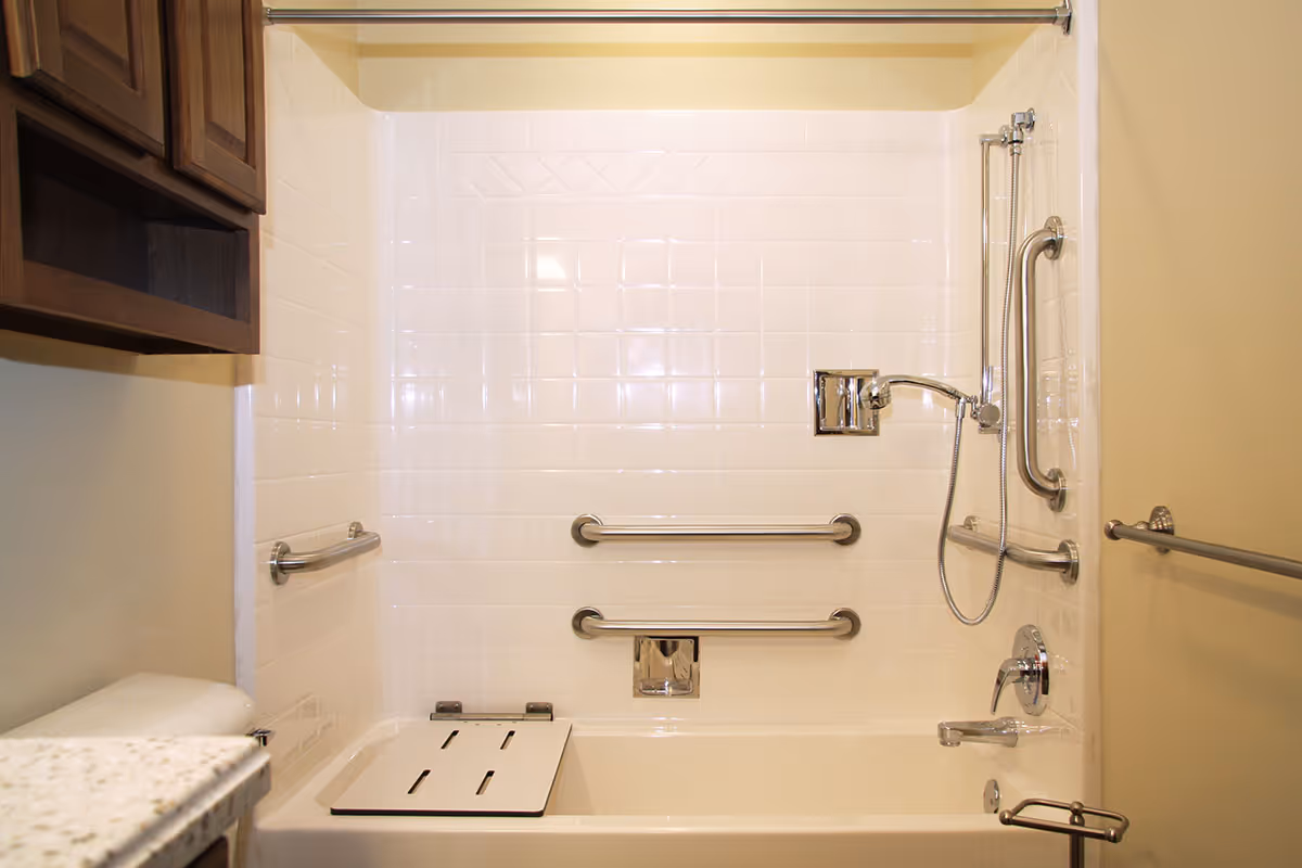 A bathroom with a white tiled bathtub and shower area equipped with multiple stainless steel grab bars and a handheld showerhead. There is a foldable shower seat attached to the bathtub. A wooden cabinet is mounted on the wall above the toilet, and a granite countertop is partially visible.