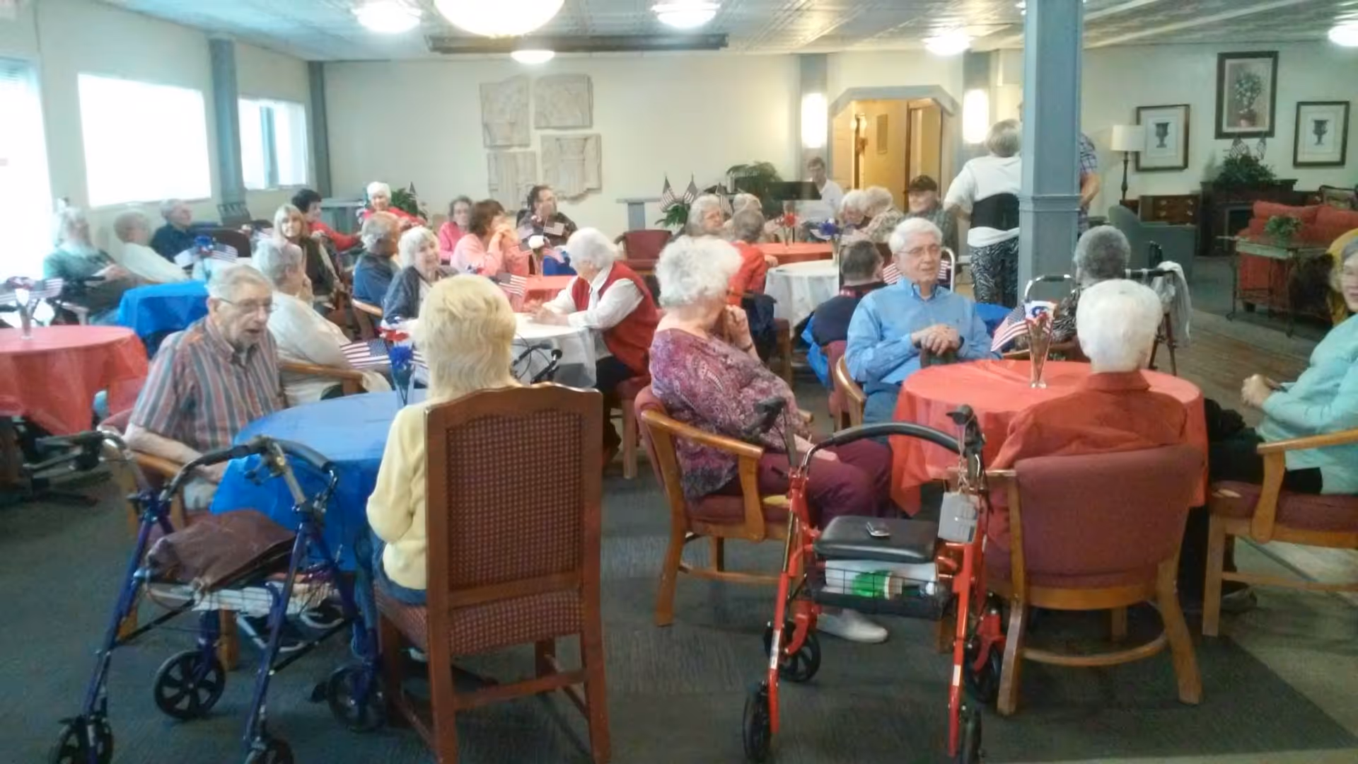 A group of elderly people sitting around tables covered with red, white, and blue tablecloths in a community room. Some have walkers nearby, and small American flags are placed on the tables. The room has framed artwork on the walls and a cozy, social atmosphere.