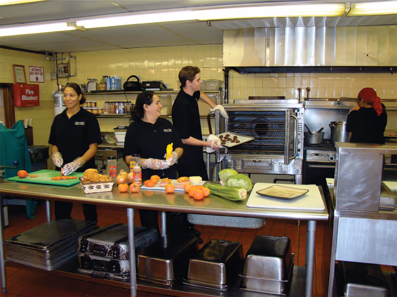 A commercial kitchen with four staff members preparing food. One person is placing a tray of cookies into an oven, two others are preparing ingredients on a stainless steel table with various vegetables and bread, and another person is working at a stove in the background. The kitchen has tiled walls and various cooking equipment.