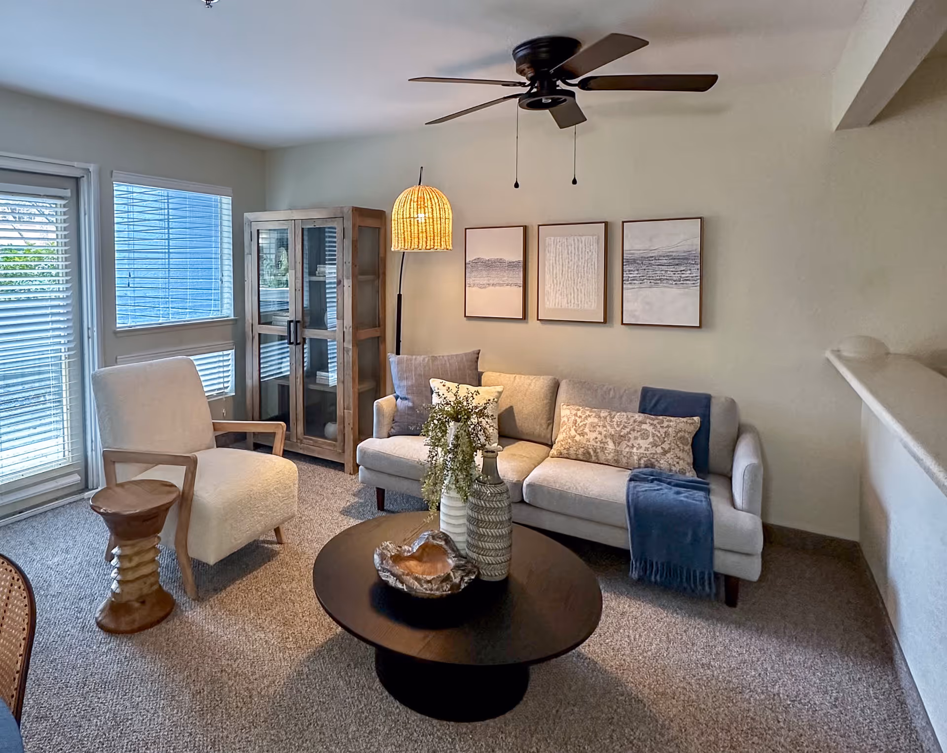A cozy living room with a beige sofa adorned with patterned and solid pillows, a blue throw blanket, a round dark wooden coffee table with decorative vases and a bowl, a cushioned armchair with wooden arms, a small wooden side table, a tall wooden cabinet with glass doors, three framed abstract artworks on the wall, a woven floor lamp, and a ceiling fan above. Large windows with blinds allow natural light into the room.