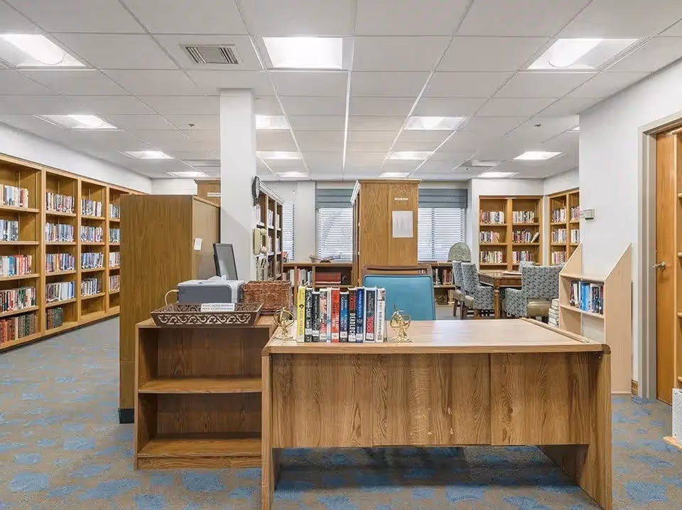 Interior view of a library room in a senior living facility with wooden bookshelves filled with books lining the walls. A wooden desk with books and decorative items is in the foreground, and several chairs and tables are visible in the background near windows with blinds.