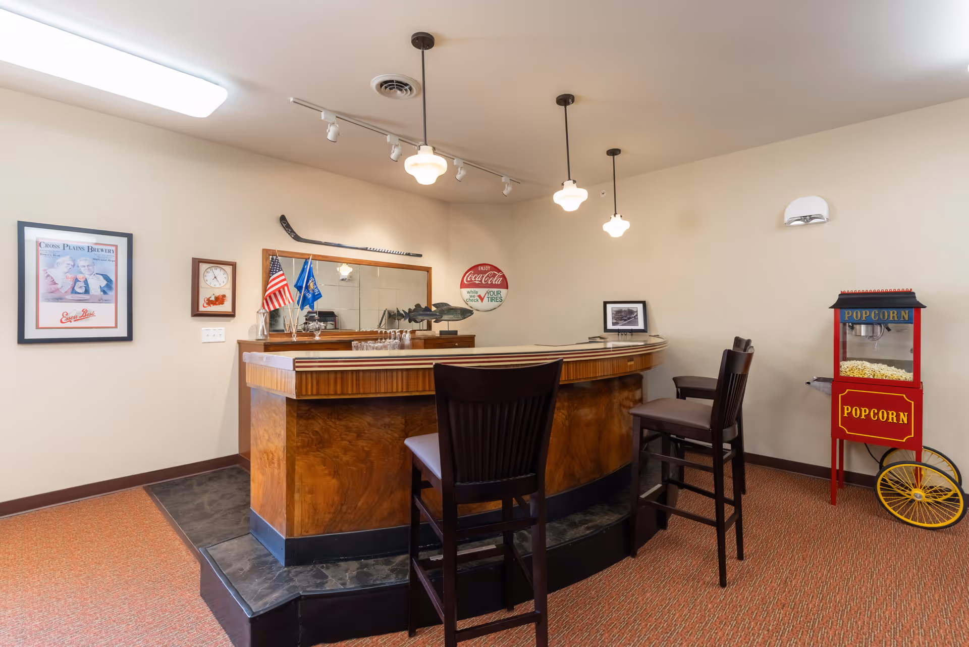 Interior view of a small bar area with a wooden counter, two high chairs, a popcorn machine on wheels, and various wall decorations including flags, a clock, a framed picture, and a vintage Coca-Cola sign.