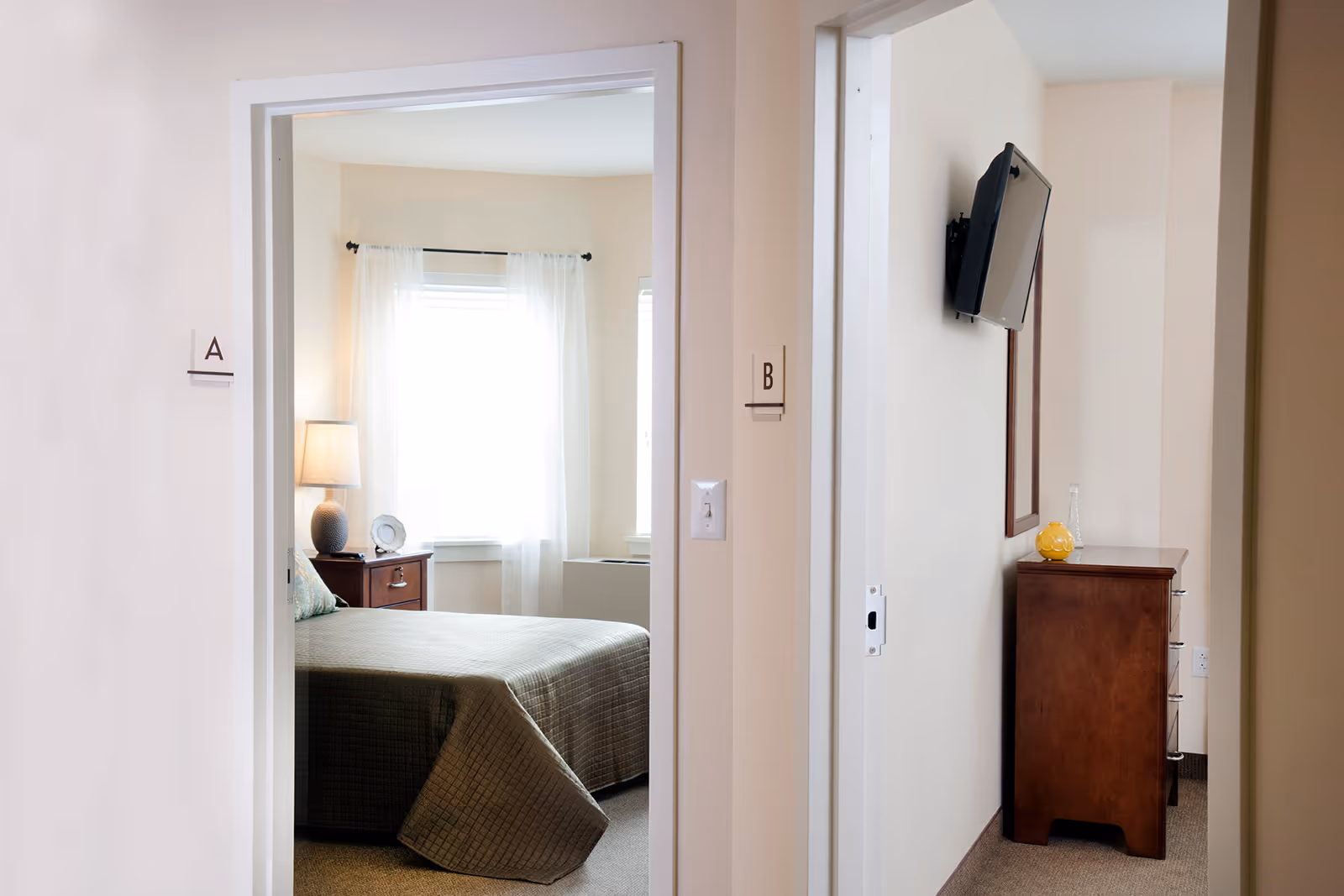 View through two adjacent doorways labeled A and B in a senior living facility. Room A shows a bedroom with a bed covered in a green quilt, a nightstand with a lamp and decorative plate, and a window with sheer white curtains. Room B shows a hallway with a mounted flat-screen TV, a wooden dresser with a yellow decorative item and a glass vase, and a mirror on the wall.