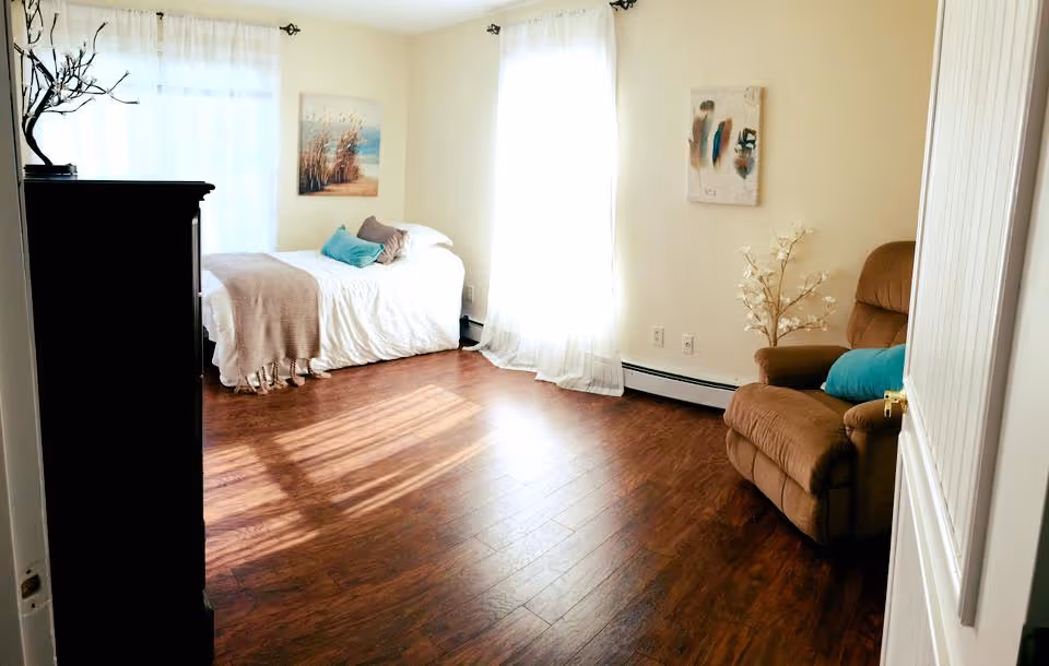 A bright and cozy bedroom with wooden flooring, a single bed with white bedding and a beige throw blanket, a brown recliner chair with a blue cushion, two large windows with sheer white curtains, a dark wooden dresser, and two pieces of wall art.