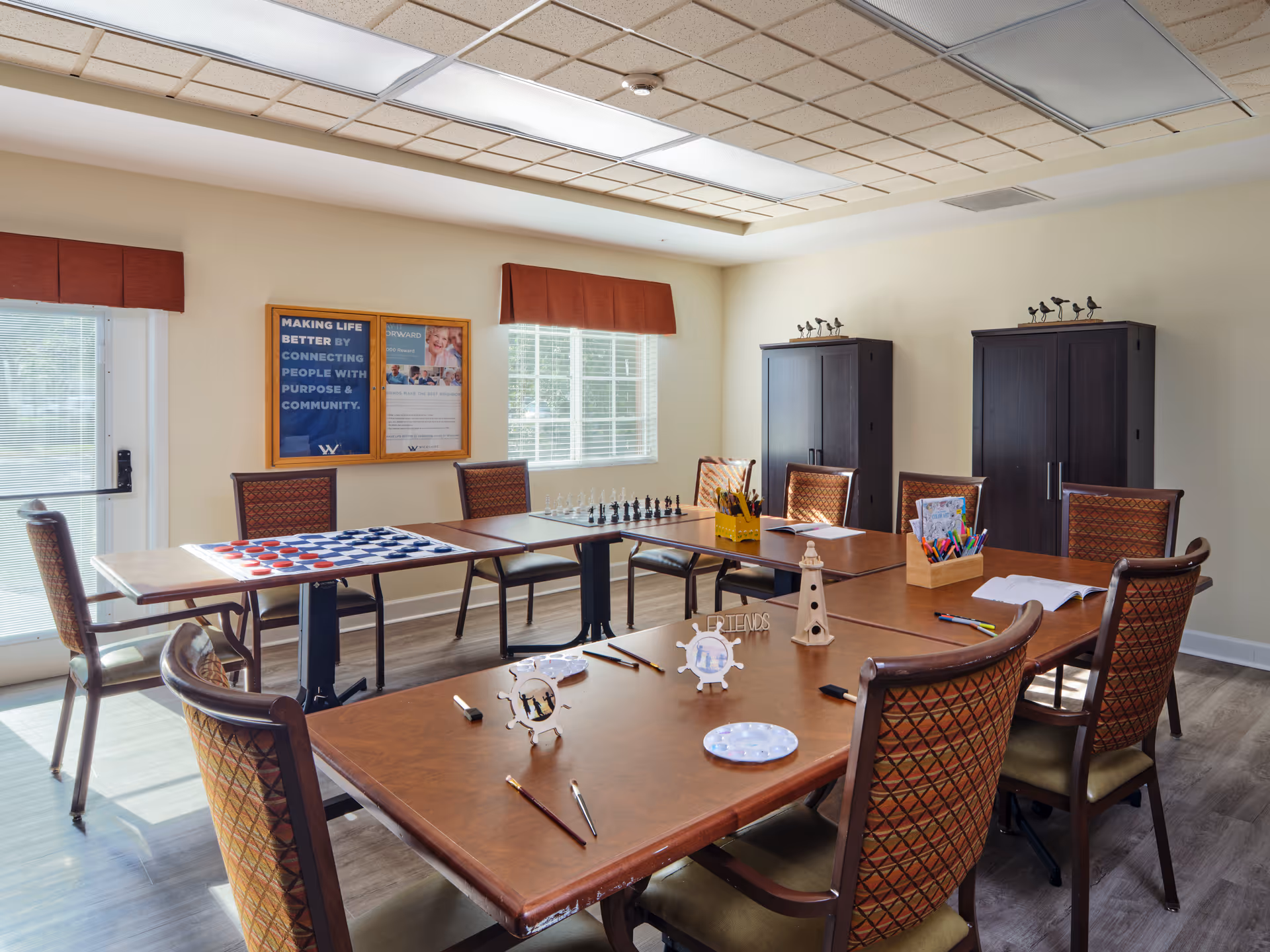 A well-lit activity room with several tables arranged in a square, surrounded by cushioned chairs. The tables have various art supplies, a chessboard, and a checkers board. Two large windows with red valances allow natural light to fill the room. On the wall, there are two framed posters, and two dark wooden cabinets stand against the far wall.
