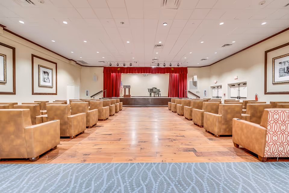 Large auditorium-style activity room with rows of brown armchairs facing a stage with red curtains and a piano.